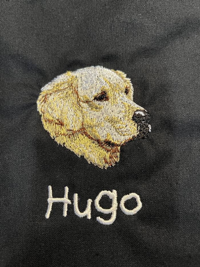 Koiran pään kuva ja nimi Hugo brodeerattuna mustaan kankaaseen
